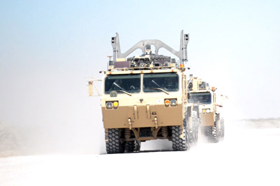 One large military transport vehicle follows another in a dusty, desert environment. 