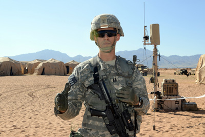 A man in a military uniform stands in a desert environment.  He has a helmet on and is holding a rifle.
