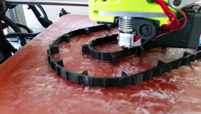 A 3D printer prints what appears to be a belt or tiny tank tracks atop a red-colored table.