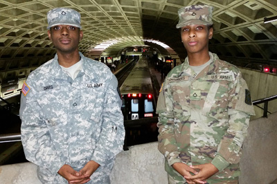 A man and a woman, both in military uniforms, stand inside a subway station.  Behind them, and one floor down, a train is in the station and picking up passengers.