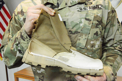 A person in a military uniform holds a military boot which has been cut in a way that reveals its cross-section.  The inside of the boot as well as the make-up of the sole is visible.