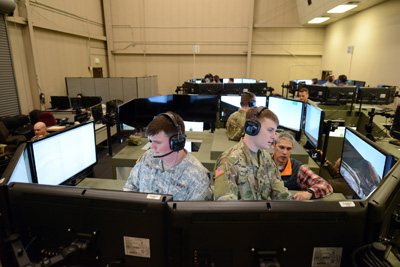 Two men in military uniforms are standing, and surrounded by a half-dozen computer monitors. A civilian man assists them. There are other similar setups in the same room.