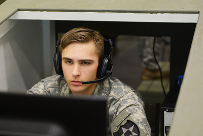 A young man in a military uniform wears a headset with a microphone. He is seated in an enclosure and is looking at a computer screen.