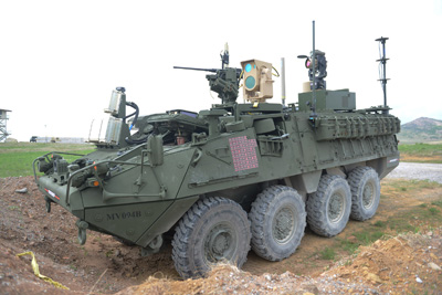 A large military vehicle sits near an earthen berm.  Atop the vehicle is a gun and a variety of optical equipment.