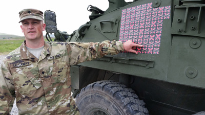 An array of stickers affixed to the side of a military vehicle indicates the number of "kills" made by the vehicle's crew.  A man in a military uniform points at the stickers.