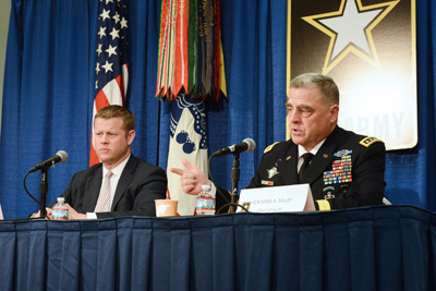 Two men, one in civilian clothing and one in a military uniform, are seated at a table.   In the rear is the logo representing the U.S. Army, the Army flag, and the American flag.