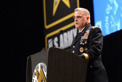 A man in military uniform stands behind a lectern.  In the rear is the logo representing the U.S. Army.