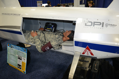 A dummy in military uniform lays on its back inside the cargo hold of a small autonomous aerial vehicle.  A medical device sits on its chest.  The words "DPI UAV Systems" and "Tribalco" appear on the side of the vehicle.