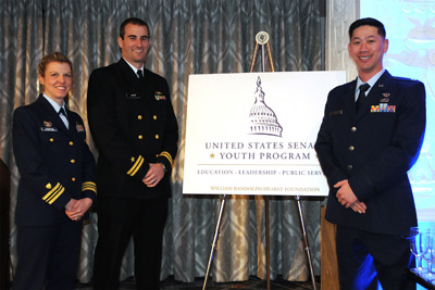 Three individuals in military uniforms stand in front of a placard that bears the words "United States Senate Youth Program."