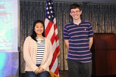 Two young people, a female and a male, stand near each other and in front of an American flag.