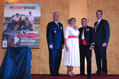 Two men in military uniforms stand alongside a man and woman in civilian clothing. To their left is a large replica of the cover of "Military Spouse" magazine on an easel. 