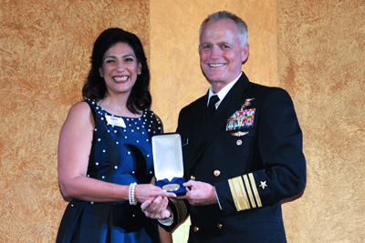 A woman in civilian clothing stands alongside a man in a military uniform.   They both hold a small box with a military medal inside.
