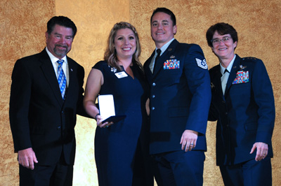 A man and woman in civilian clothing stand alongside a man and woman in military clothing.  The women in civilian clothing holds a box with a military medal inside.