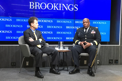 Two men, one in a military uniform, are seated near each other on a stage.  Behind them, a display screen reads "Brookings."