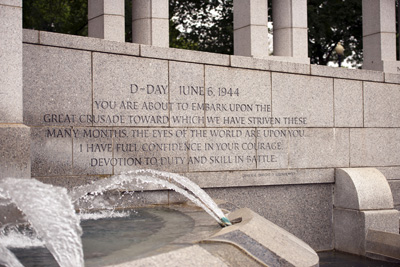A quote about D-Day is engraved in a stone wall.  A fountain sprays water.