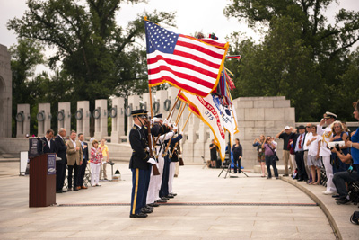 Uniformed military personnel stand outdoors in a line and carry flags, amidst fountains and ornate stonework. Others are standing and saluting.