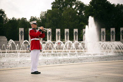 A man in a red uniform plays a trumpet outdoors, amidst fountains and ornate stonework.