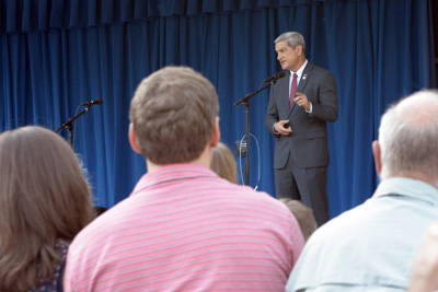 A man in a suit stands behind a microphone on a stage. Others are seated in front of him.