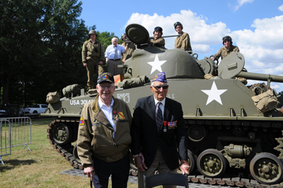 Two elderly men stand together outdoors in front of a military tank. Several other men stand on the tank.