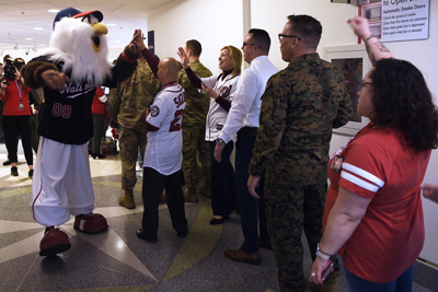 Indoors, a man in an eagle mascot costume, emblazoned with the word "Nationals," greets individuals lined up against a wall.