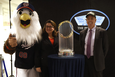 A person dressed like a large eagle stands next to a woman, a man, and a trophy. Behind them the Pentagon logo is emblazoned on the wall.