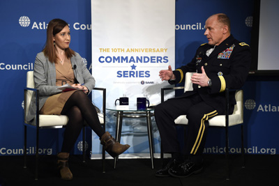 A woman in a business suit and a man in uniform are seated and facing each other on a stage. A banner reads "The 10th Anniversary Commanders Series." The words "Atlantic Council" appear on the wall.