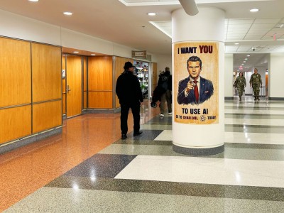 In a large public area, individuals walk past a column that features a poster which features a man pointing, and the words "I want you to use AI."
