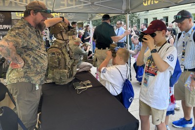 Two boys look through goggles, while outside and under a tent.  A mannequin wearing a military uniform also wears the goggles. An older man watches.