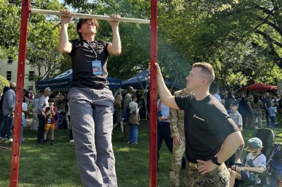 A young man does pull-ups on a bar. A Marine watches him.  They are outdoors.
